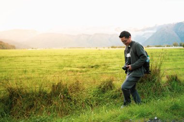 Genç Asyalı fotoğrafçı elinde kamerayla yürüyüp Yeni Zelanda 'daki ünlü yer olan Matheson Gölü' nün arkasındaki çitin arkasındaki güzel yeşil manzarayı yakalamak için iyi bir yer arıyor..