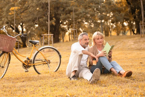 Happy caucasian mature couple relax and enjoy the afternoon autumn sun in the park with coffee and book after riding a bicycle, concept relax lifestyle, carefree retirement lifestyle, couple love time