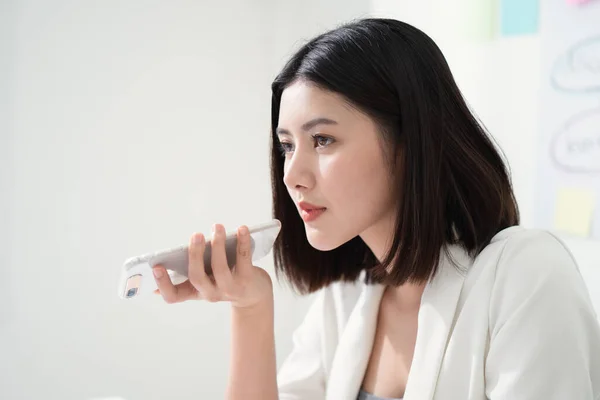 Young beautiful asian business woman using speech recognition to communication on mobile phone while sitting in the white office, voice or speech recognition is the powerful  for new age technology.