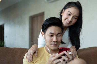 Akıllı telefonu güzel bir kadının arkasında müzik dinlemek için kullanan bir adam oturma odasında çekici bir Asyalı çift mutlu genç adam ve kadın sıradan bir yaşam tarzında. Sevgili karı koca ilişkisi..