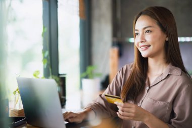 Bilgisayarla online alışveriş yapmak için elinde kredi kartıyla gülümseyen genç bir Asyalı kadın..