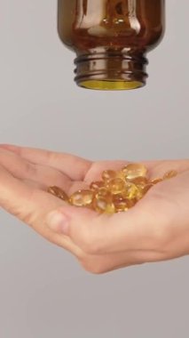 Kahverengi bir şişeden dökülen altın yumuşak kapsüllerle dolu bir elin dikey yakın çekimi. Vitamin, balık yağı, omega 3 takviyesi, sağlık hizmetleri ve günlük sağlık rutini kavramı.
