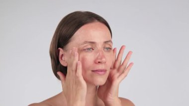 Yüz serumu ya da yüz yağını açık arkaplana sürerken yüzüne nazikçe dokunan bir kadının portresi. Sıvılaştırıcı cilt bakımı ve fotokopi alanı ile kendine bakım kavramı.