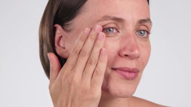 Gülümseyen bir kadının, arka planda yanağına nemlendirici sürerken yakın çekimi. El yapımı cilt bakımı ürünü, SPF ile makyaj temeli. Hassas cilt için günlük yüz bakımı.