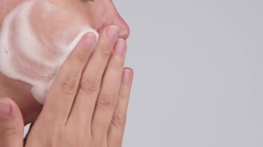 Arka planda temizleyici köpükle yüzünü yıkayan tanınmayan bir kadının yakın çekimi. Yüz derisi rutini sırasında yanaktaki köpüğe kopyalama alanı ile el masajı yapmak..