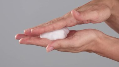 Stüdyoda arka planda, avuçların arasına beyaz köpük geren bir kadının yakın çekimi. Cilt bakımı, vücut bakımı ve hijyen rutini sırasında ürün dokusu tanıtımı.