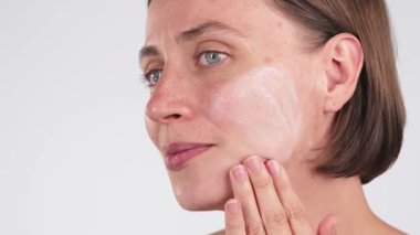 Gülümseyen bir kadının, tarafsız bir arkaplanda yanağına nemlendirici krem sürerken yakın çekimi. Günlük yüz bakımı, kolajen tedavisi, cilt bakımı, cilt bakımı....