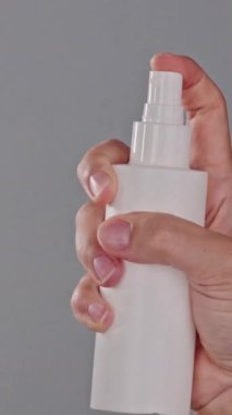 Tarafsız bir arkaplanda beyaz bir şişeden ince bir sis püskürten bir elin dikey yakın çekimi. Saç spreyi ya da ısı koruması kullanılıyor. Saç bakım ürünü reklamcılığı konsepti.