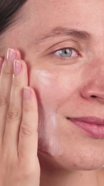 Gülümseyen bir kadının yanağına nemlendirici sürerken dikey yakın çekimi. El yapımı cilt bakımı ürünü, SPF ile makyaj temeli. Hassas cilt için günlük yüz bakımı.