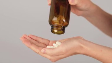 Kahverengi cam bir şişeden dökülen beyaz tabletlerle dolu bir elin yakın çekimi. İlaçlar, multivitaminler, ağrı kesici ve günlük sağlık hizmetleri rutini.