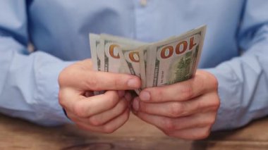 Erkek ellerinin 100 dolarlık banknotları saydığı yakın çekim. Mavi gömlekli adam para sayıyor, Amerikan parası. Gelir, maaş veya tasarruf planlaması için mali konsept.