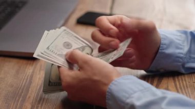 Mavi gömlekli adam ahşap masada 100 dolar sayıyor. Adam para sayıyor. Gelir, tasarruf ya da bütçe planlaması için finansal konsept. Kişisel finans, iş veya yatırım içeriği için ideal.