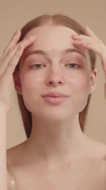 Serum kremi sürdükten sonra güzel yüzüne masaj yapan genç bir kadın kameraya bakıp doğal güzelliğin, temiz, nemli ve sağlıklı cilt bakımının tadını çıkarıyor. Dikey video