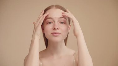 Serum kremi sürdükten sonra güzel yüzüne masaj yapan çekici beyaz kadın kameraya bakıp doğal güzelliğin, temiz, nemli ve sağlıklı cilt bakımının tadını çıkarıyor.