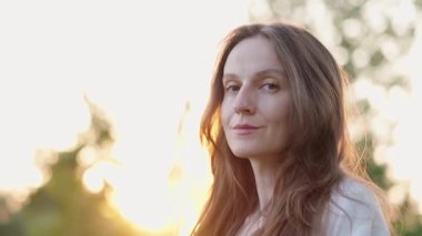 Uzun, dalgalı saçlı gülümseyen esmer kadının portresi, açık havada gün ışığında kameraya bakıyor. Ağır çekim. Düşük açı görünümü.