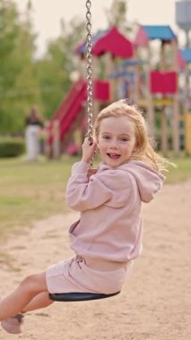 Heyecanlı kız salıncakta sallanmaktan zevk alıyor. Mutlu çocuk oyun alanında eğleniyor, neşeli bir şekilde gülümsüyor ve açık havada eğleniyor..