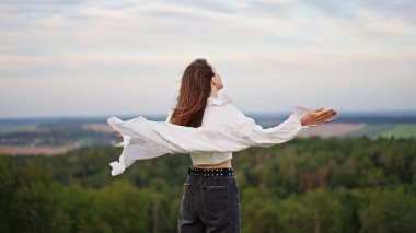 Genç bir kadın kaldırılmış kollarıyla tepenin üstünde, rüzgarı hissediyor saçları ve gömleğiyle.