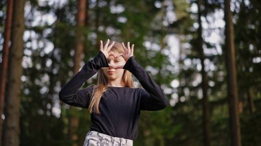 Genç kız Alfa dışarıda parkta duruyor, elleriyle kalp şekli oluşturuyor, kameraya bakıyor, nazikçe gülümsüyor ve sevgi yayıyor, pozitiflik..