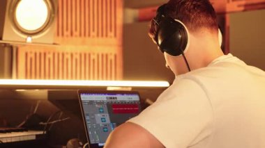Genç Yapımcı Dizüstü bilgisayarda Müzik Parçası Düzenliyor, Kulaklıklar açık. Stüdyo Sahnesi Yaratıcı Ses Tasarımı, Modern Müzik Yapımı, Sürece Odaklanma Gösteriyor.