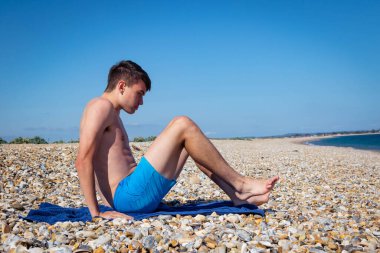 18 yaşında, beyaz, üstsüz bir çocuk taşlı bir sahilde oturuyor.