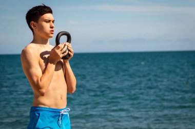Plajda kettlebell ağırlığıyla egzersiz yapan 18 yaşında beyaz gömleksiz bir genç.