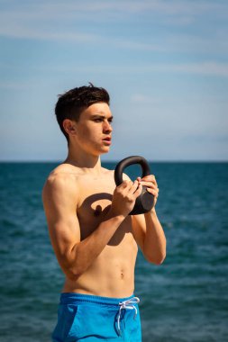 Plajda kettlebell ağırlığıyla egzersiz yapan 18 yaşında beyaz gömleksiz bir genç.