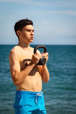 Plajda kettlebell ağırlığıyla egzersiz yapan 18 yaşında beyaz gömleksiz bir genç.