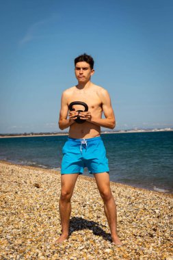 Plajda kettlebell ağırlığıyla egzersiz yapan 18 yaşında beyaz gömleksiz bir genç.