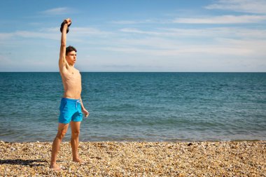 18 yaşında, beyaz tişörtsüz mayo giyen genç bir çocuk. Fotokopi odasıyla kettlebell ağırlığında egzersiz yapıyor.
