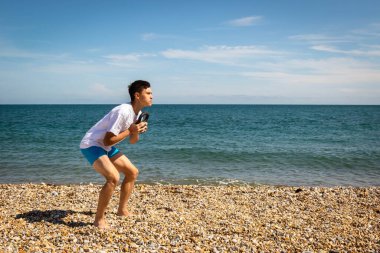 18 yaşında, beyaz bir çocuk plajda, elinde bir çaydanlık ağırlığıyla fotokopi uzayı ile egzersiz yapıyor.