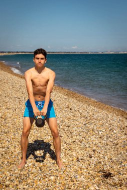 Plajda kettlebell ağırlığıyla egzersiz yapan 18 yaşında beyaz gömleksiz bir genç.