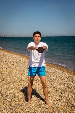 Plajda kettlebell ağırlığıyla egzersiz yapan 18 yaşında beyaz bir çocuk.