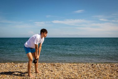 Plajda kettlebell ağırlığıyla egzersiz yapan 18 yaşında beyaz bir çocuk.
