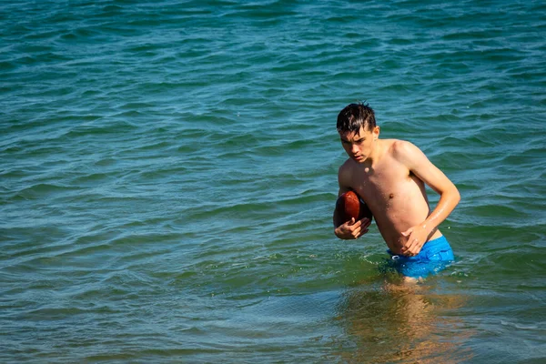 一名18岁的白种人少年穿着泳衣在海里与美式足球玩耍 — 图库照片