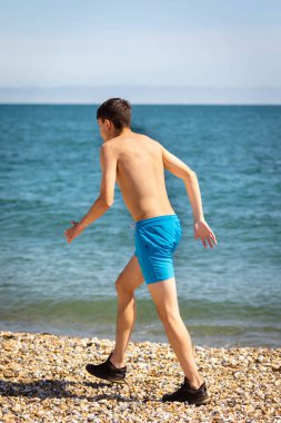 Denizin kenarında sahilde yürüyen 18 yaşında, beyaz tişörtsüz genç bir çocuk.