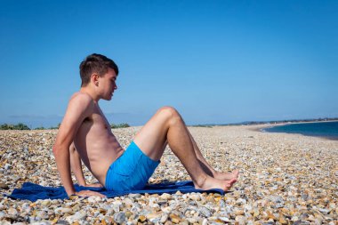 18 yaşında, beyaz, üstsüz bir çocuk taşlı bir sahilde oturuyor.