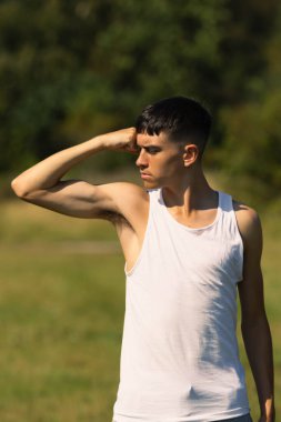 18 yaşında kolsuz tişörtlü bir genç yaz günü kol kaslarını esnetiyor.