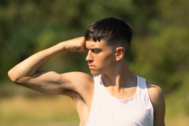18 yaşında kolsuz tişörtlü bir genç yaz günü kol kaslarını esnetiyor.