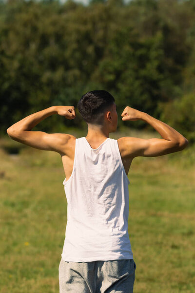 An 18 year old teen boy in a sleeveless top on a late summer's day, flexing his arm muscles