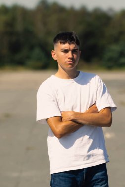 18 yaşında, açık havada beyaz tişörtlü ve kolları katlanmış kot pantolonlu bir çocuk.