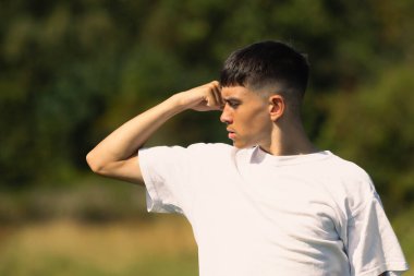 18 yaşında, beyaz tişörtlü, kollarından birini esnetiyor.