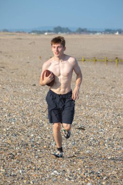 Genç bir çocuk, sıcak bir yaz gününde kumsalda elinde Amerikan futbolu topuyla kameraya doğru koşuyor.