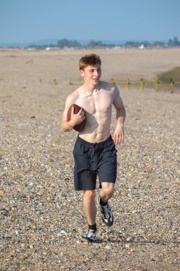 Genç bir çocuk, sıcak bir yaz gününde kumsalda elinde Amerikan futbolu topuyla kameraya doğru koşuyor.