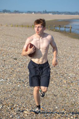 Genç bir çocuk, sıcak bir yaz gününde kumsalda elinde Amerikan futbolu topuyla kameraya doğru koşuyor.