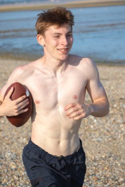 Genç bir çocuk, sıcak bir yaz gününde kumsalda elinde Amerikan futbolu topuyla kameraya doğru koşuyor.