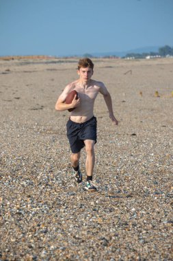 Genç bir çocuk, sıcak bir yaz gününde kumsalda elinde Amerikan futbolu topuyla kameraya doğru koşuyor.