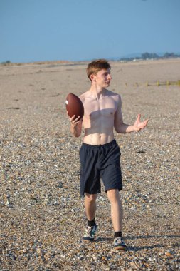 Genç bir çocuk, sıcak bir yaz gününde kumsalda elinde Amerikan futbolu topuyla kameraya doğru yürüyor.