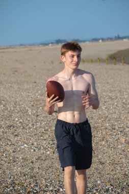 Genç bir çocuk, sıcak bir yaz gününde kumsalda elinde Amerikan futbolu topuyla kameraya doğru yürüyor.