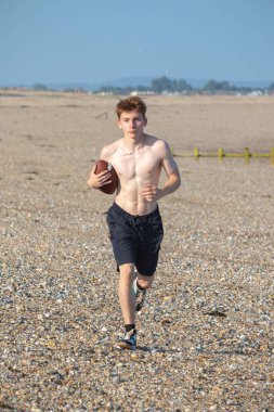 Genç bir çocuk, sıcak bir yaz gününde kumsalda elinde Amerikan futbolu topuyla kameraya doğru koşuyor.