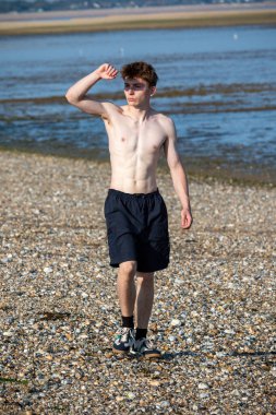 Teenage boy walking towards the camera and shielding his eyes from the sun, along a beach on warm summer's day